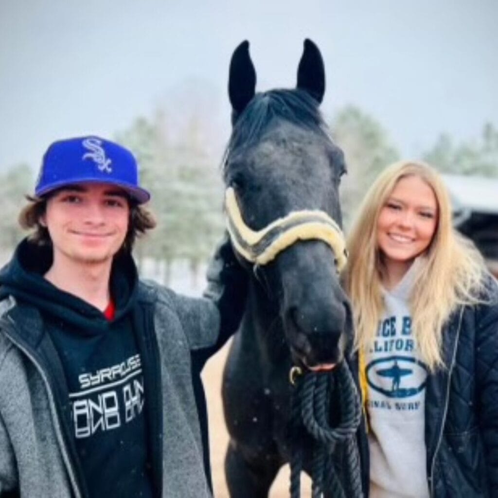 Two people standing outdoors with a black horse, smiling at the camera. One person is wearing a dark hoodie and a blue baseball cap, and the other a light hoodie with visible logos. The horse has a yellow halter on.