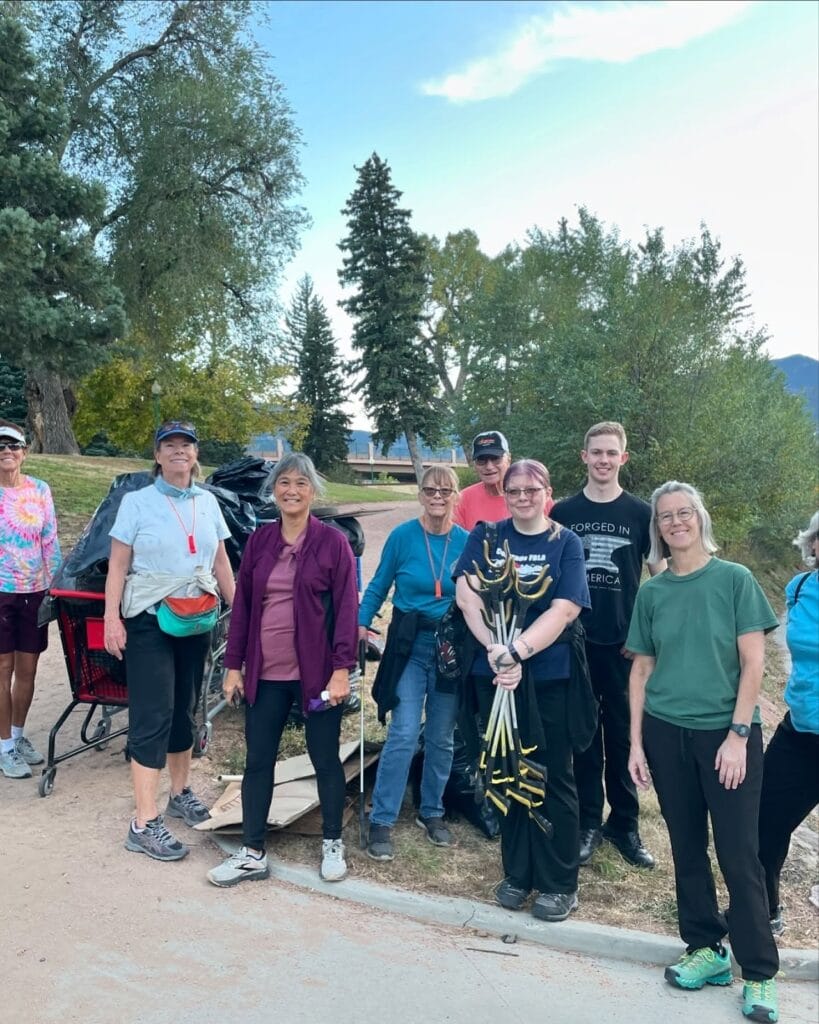 Group of individuals participating in a community clean-up event, standing outdoors with trash bags and cleanup gear, in a park with trees and mountains in the background.