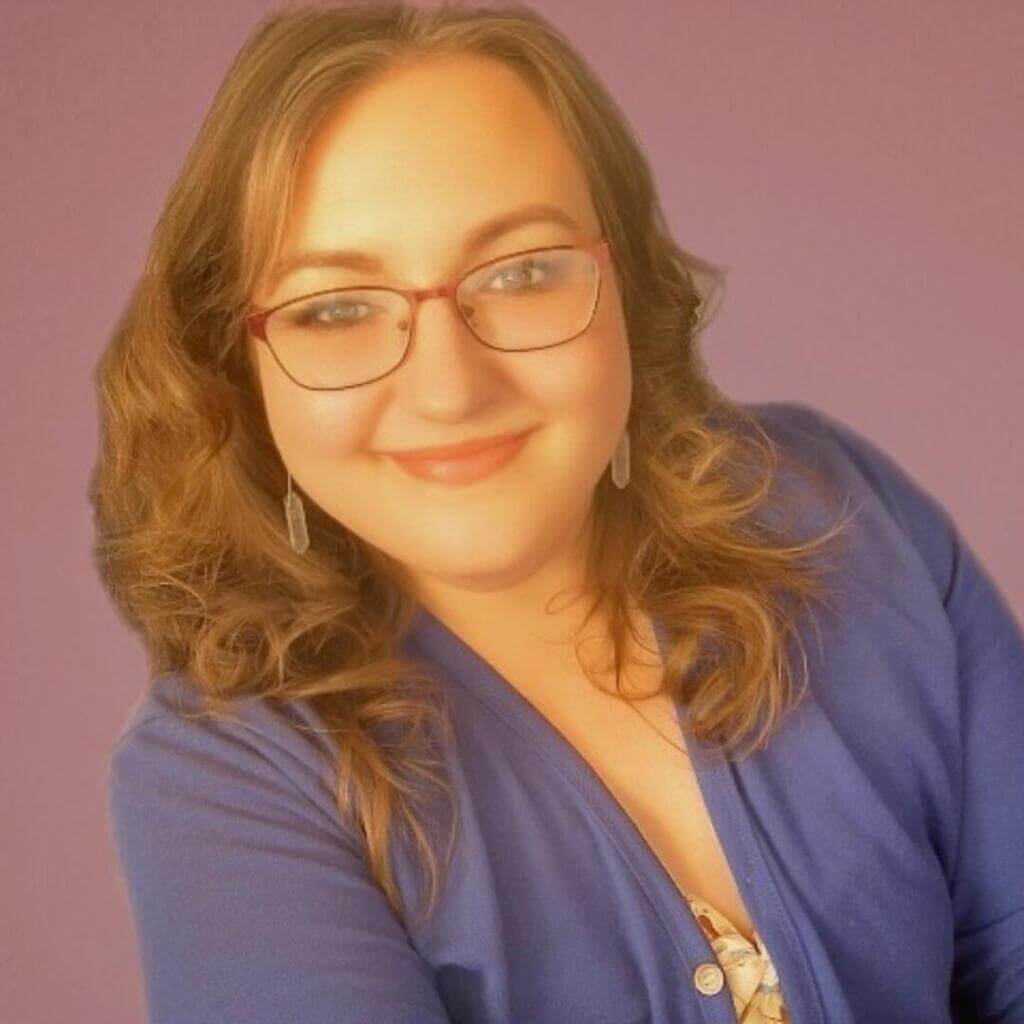 A woman with wavy light brown hair, glasses, and a confident smile poses in front of a purple background, wearing a blue top.

