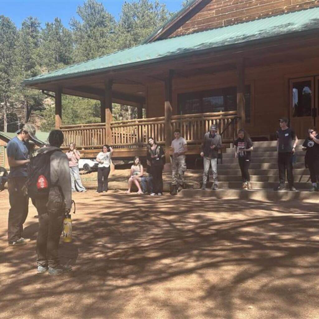 A group of young people sits together on logs and benches in a shaded forest area, talking and laughing casually near a wooden cabin.

