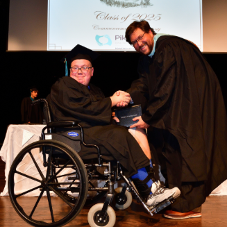 A graduate in a wheelchair receives a diploma from a man in a gown. Both smile onstage during a graduation ceremony, under a "Class of 2023" sign.