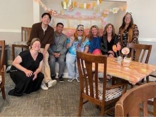 A group of seven people, smiling and seated, in a decorated room with floral and pastel garlands. The atmosphere is festive and joyful.
