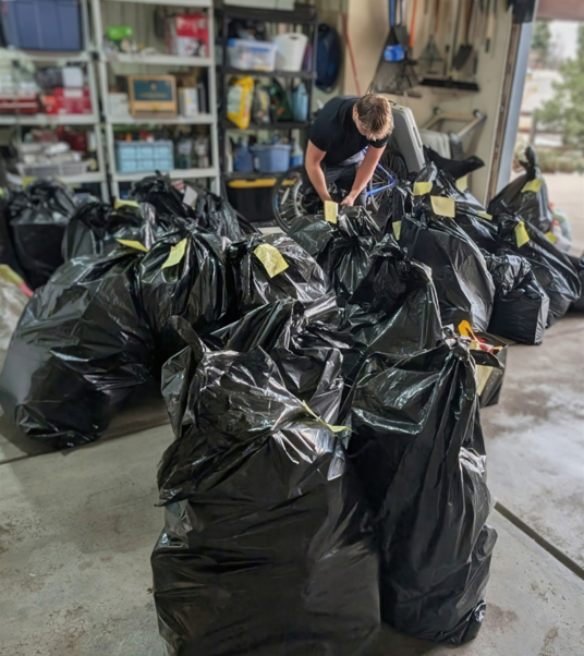 A person bends over a large group of filled black trash bags in a garage, attaching or checking labels on the bags. Shelving with storage items lines the back wall.