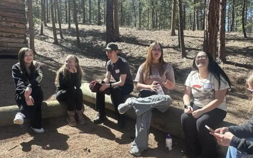 A group of people stands in a semi-circle outside a wooden cabin surrounded by tall pine trees. They appear to be listening to a speaker or guide in a sunny, wooded area.