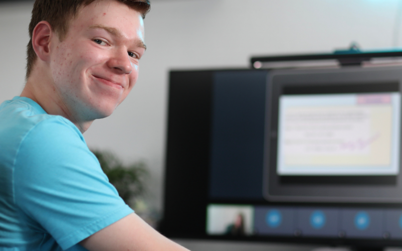 A person in a blue shirt sits at a desk participating in an online class or meeting, with a large monitor displaying a digital lesson or presentation in the background.