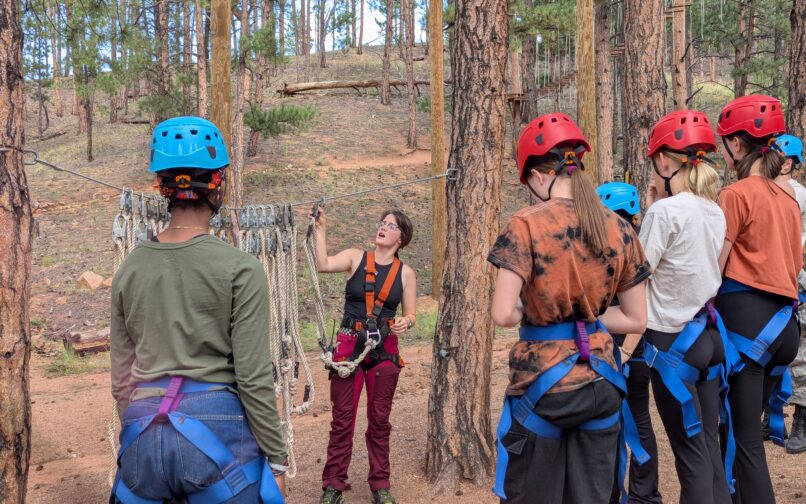 A group of individuals wearing helmets and harnesses stand among tall pine trees while listening to an instructor demonstrate ropes and climbing equipment at an outdoor adventure course.
