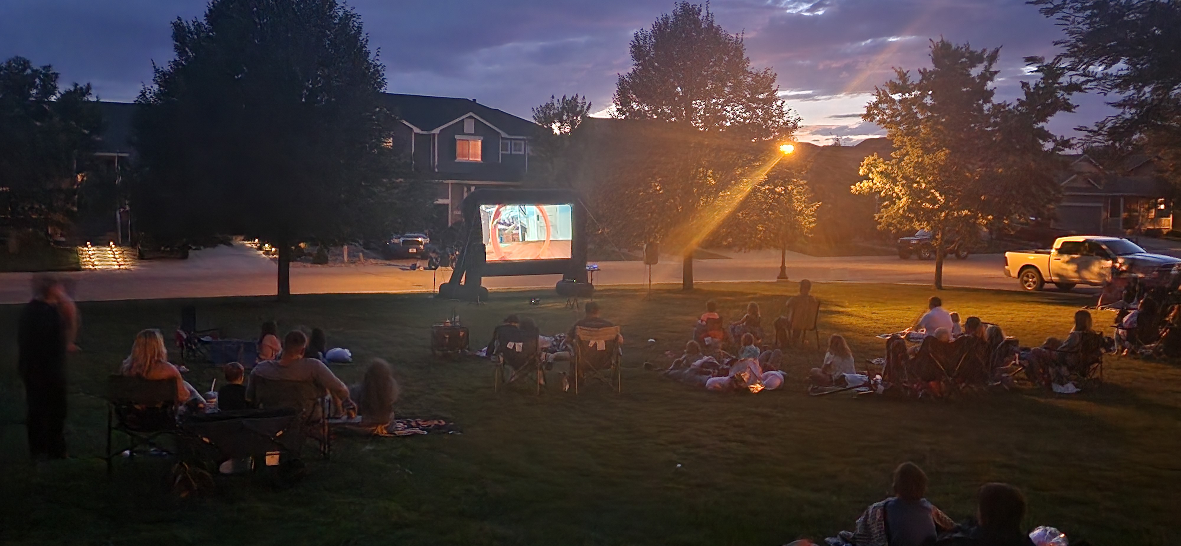 Families and community members sit on blankets and lawn chairs in a grassy park at dusk, watching a movie projected on a large outdoor screen as streetlights and nearby houses glow in the background.